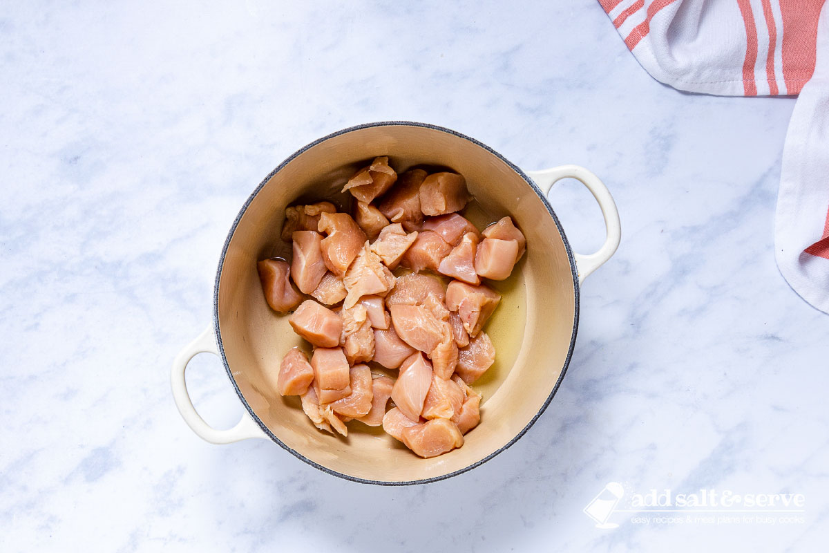 Enameled Dutch oven with oil and uncooked chopped chicken breast.