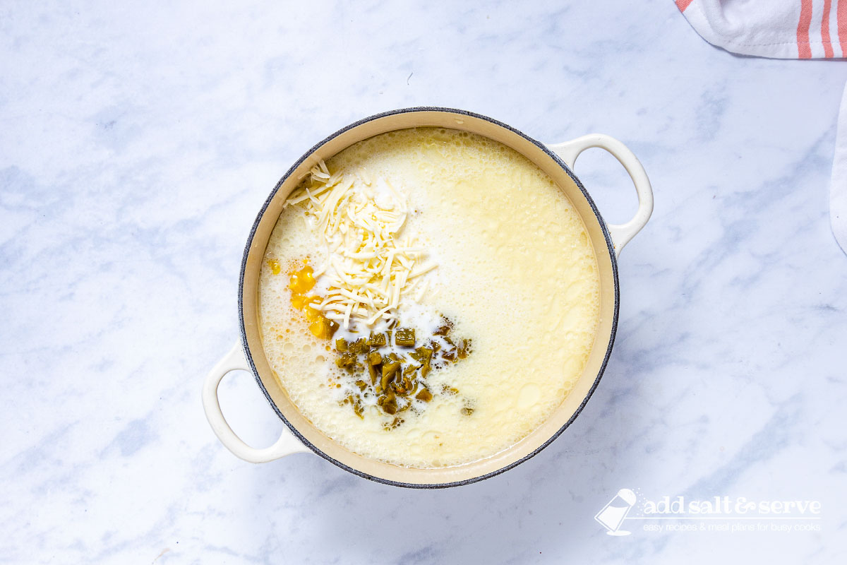 Enameled Dutch oven with ingredients for Southwest Chicken Corn Chowder after chicken and garlic has been sautéed and the following ingredients have been added: creamed corn, diced green chilies, half and half, chicken broth, and Monterey Jack cheese.