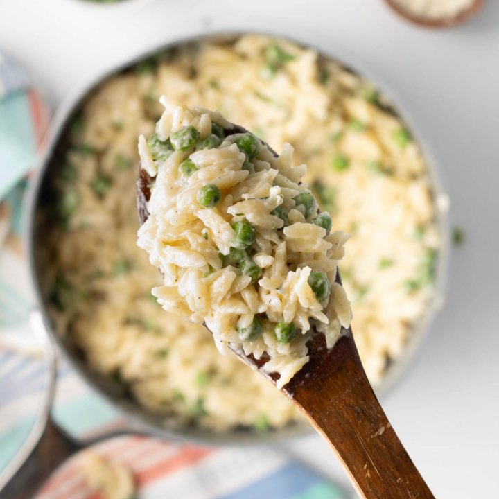 Serving of Orzo Alfredo with peas on a wooden spoon above the pot.