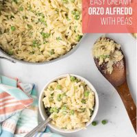 a bowl of Orzo Alfredo with peas with a fork beside the pot of the dish with a wooden spoon laying beside it.