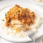 Plated slow cooker Thai Peanut Chicken over rice garnished with chopped peanuts and cilantro with a fork cutting into the chicken showing white breast meat.