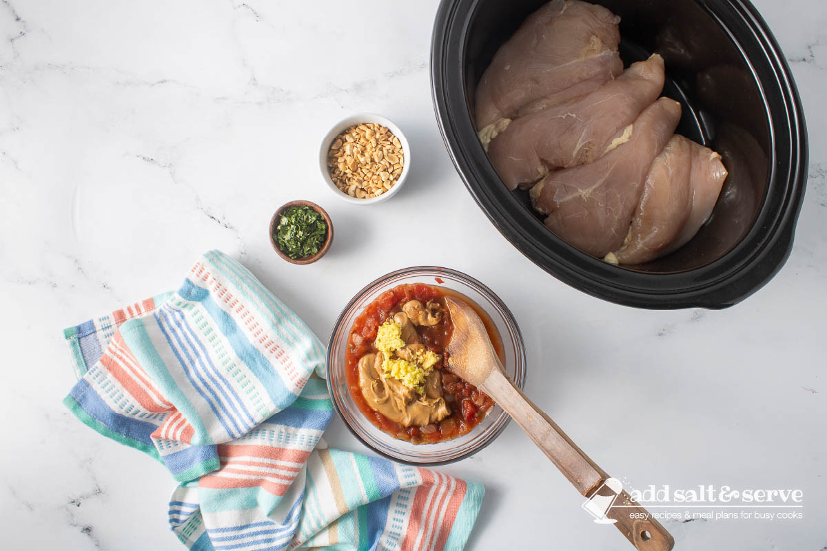 Uncooked chicken breasts in a slow cooker and small bowls of chopped peanuts and cilantro around a bowl of peanut butter, salsa, soy sauce, grated ginger, and lime juice before mixing.