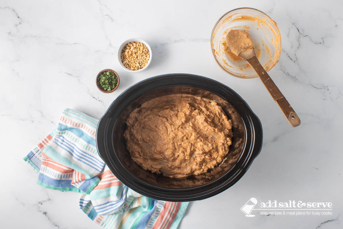 A slow cooker with chicken covered in a peanut sauce before cooking.