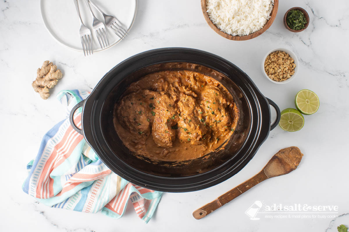Cooked Thai Peanut Chicken in a slow cooker surrounded by rice, chopped peanuts, cilantro, ginger, a halved lime, and a stack of empty plates with forks.