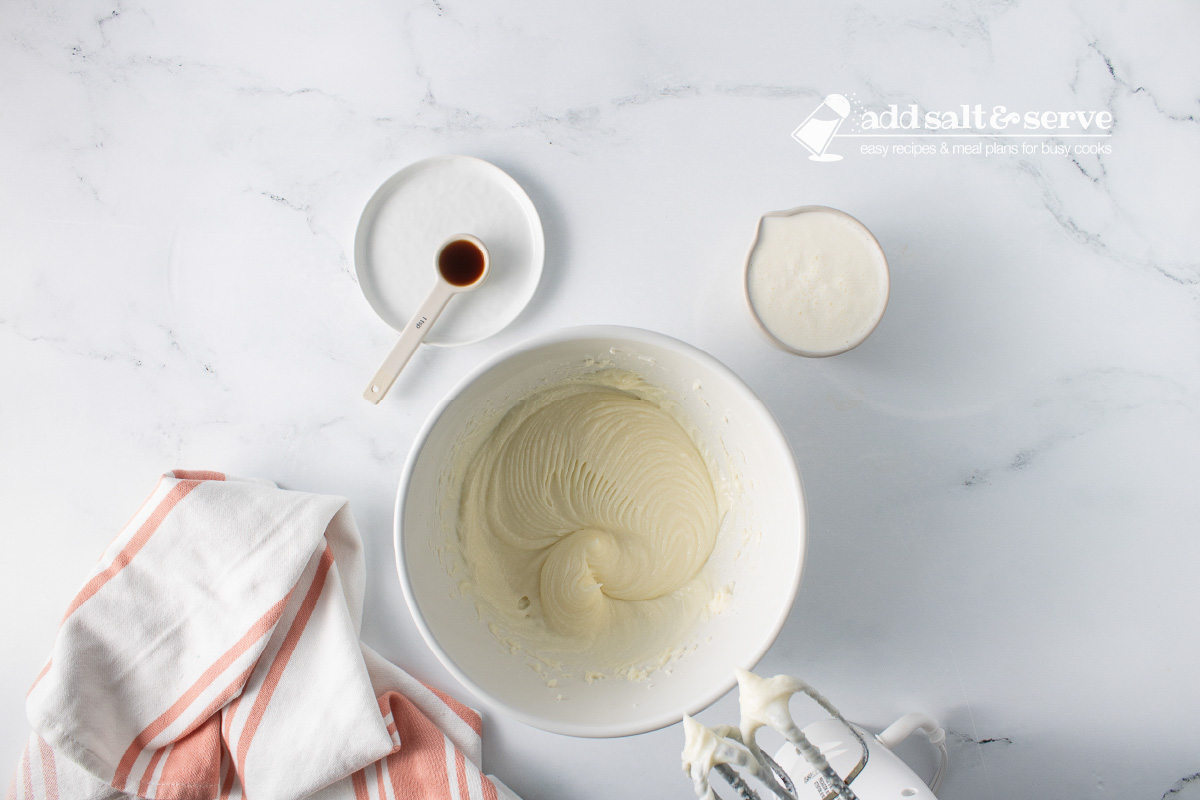 White mixing bowl with a whipped cream cheese and sugar mixture with dishes to the side with vanilla extract and heavy cream.