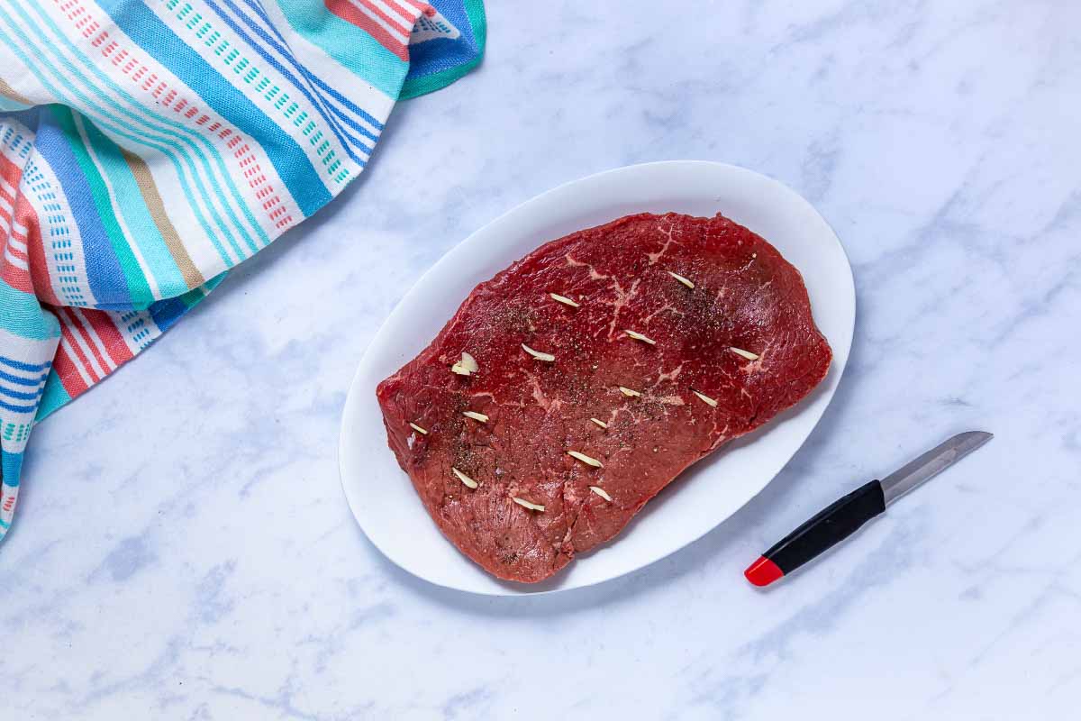 Uncooked London broil roast seasoned with salt and pepper with sliced garlic in slits cut into the roast.