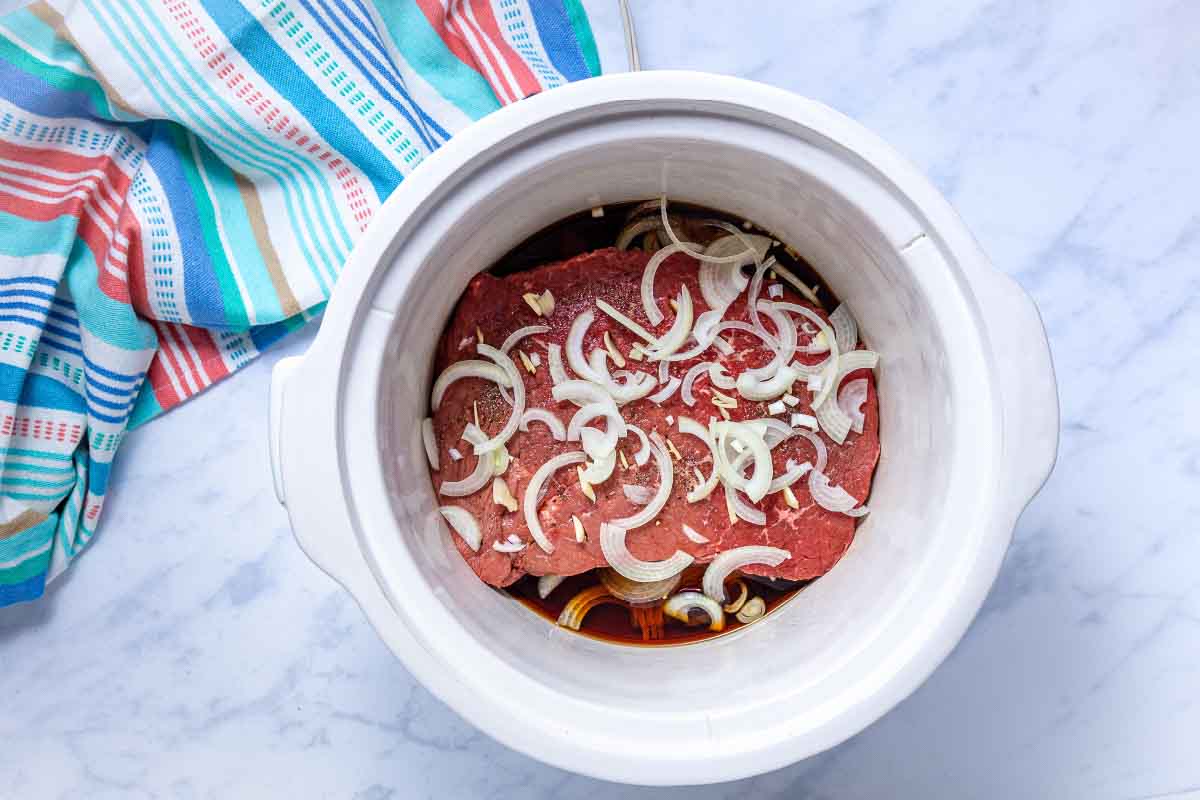 A crockpot with layers of sliced onions and seasoned uncooked London broil roast covered in soy sauce and water.