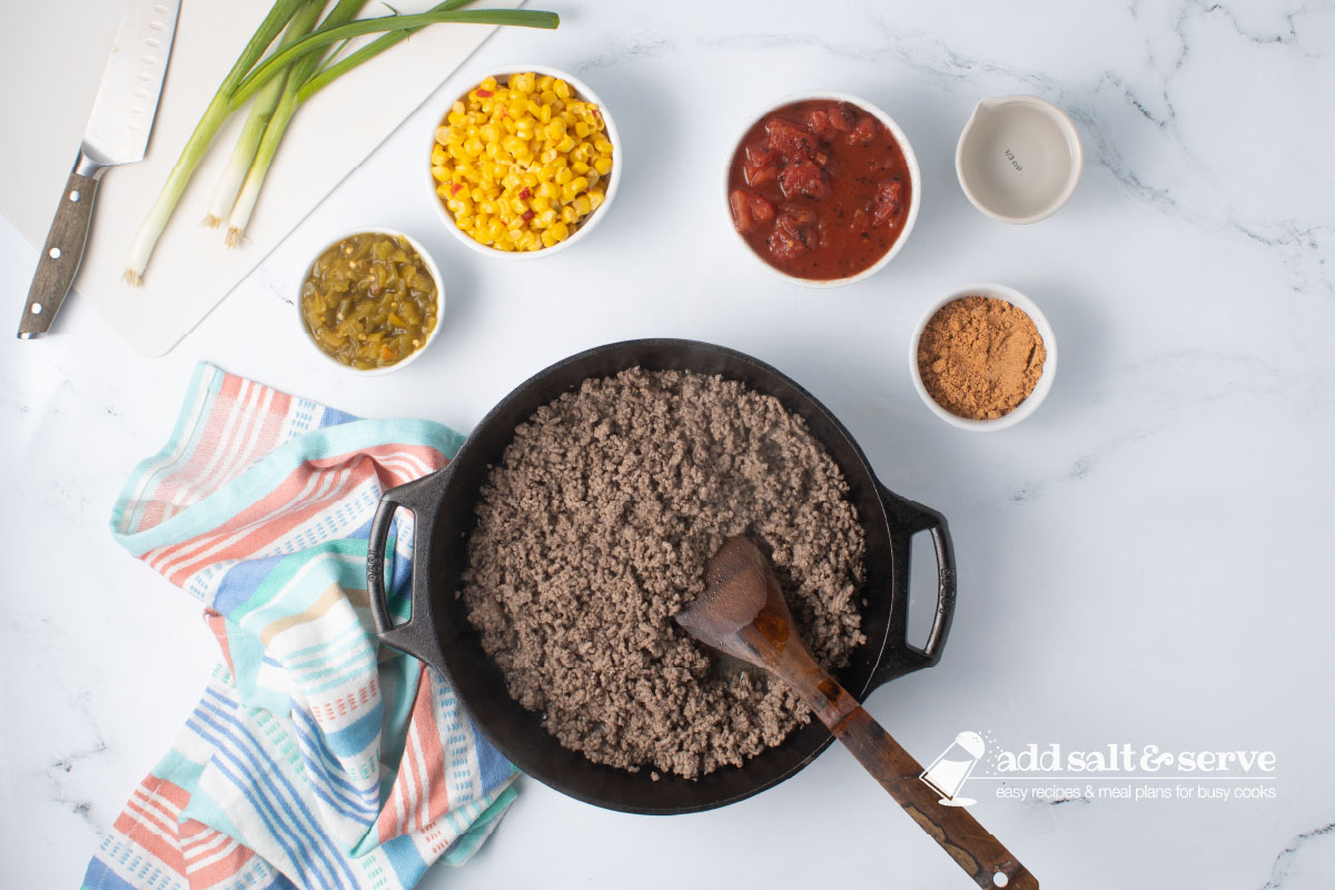 Cast iron skillet with cooked ground beef wtih a wooden spoon surrounded by bowls of green chilis, mexi-corn, fire-roasted diced tomatoes, taco seasoning, and water.