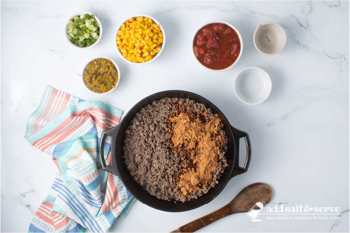 Cast iron skillet with cooked ground beef and taco seasoning surrounded by bowls with mexi-corn, green chilis, fire-roasted diced tomatoes, water, and chopped green onions and a wooden spoon.