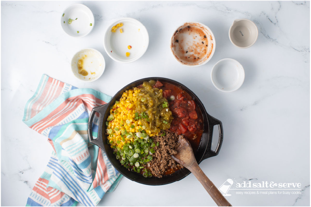 Cast iron skillet with seasoned cooked ground beef, mexi-corn, green chilis, fire-raosted diced tomatoes, and chopped green onions with a wooden spoon.