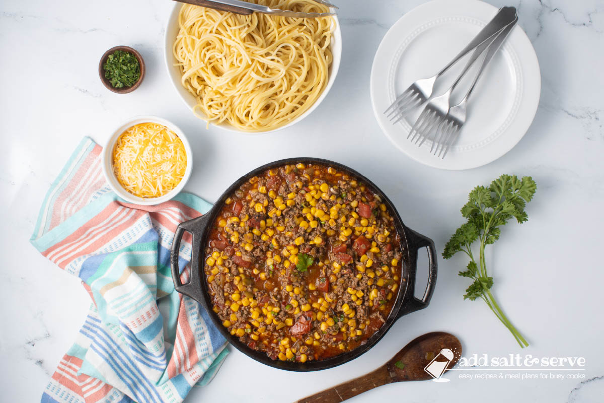 Cast iron skillet of Mexican Spaghetti sauce, a dish of shredded cheddar cheese, and a bowl of cooked spaghetti on a counter next to fresh parsley.