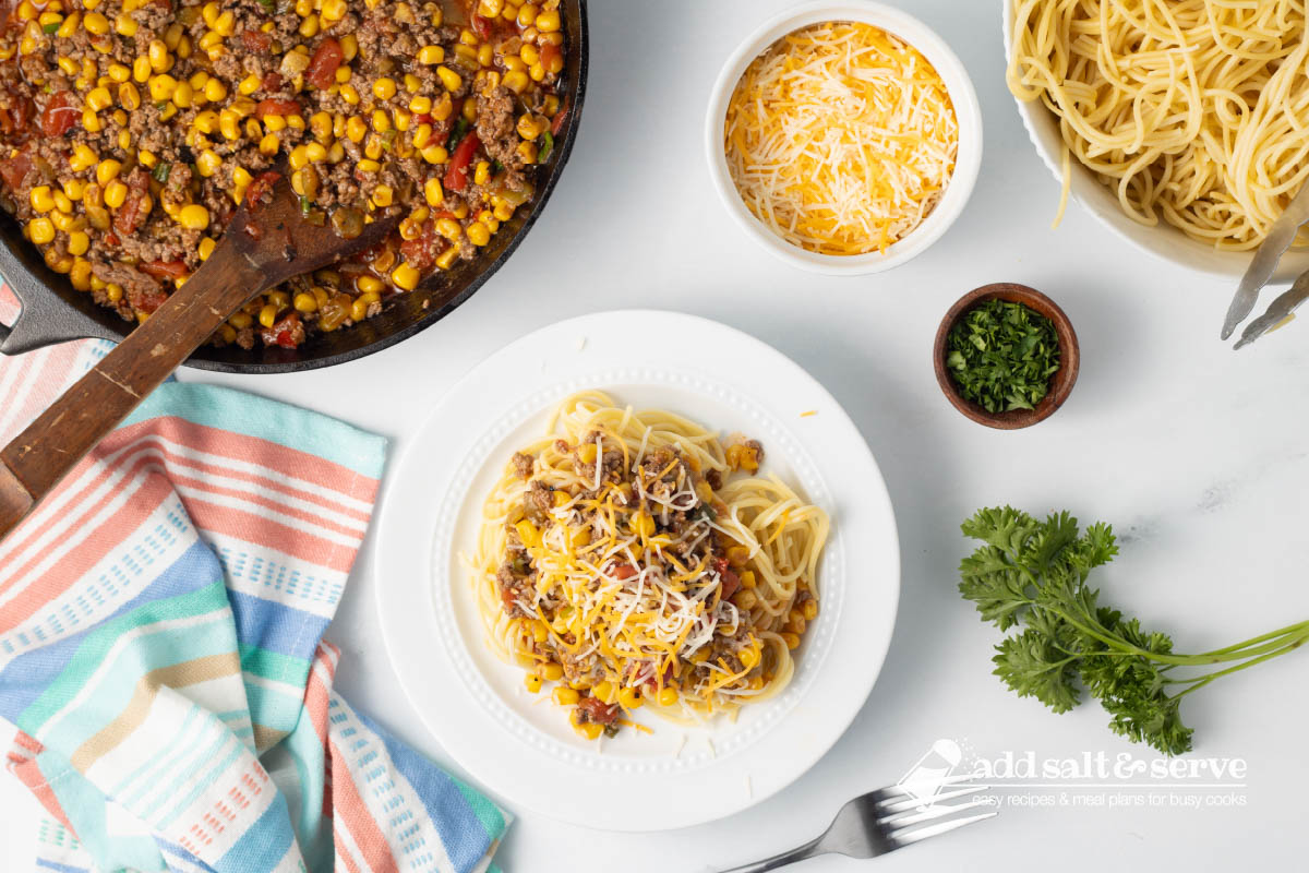 Plate of Mexican Spaghetti topped with shredded cheese beside a bowl of cooked spaghetti, a pot of Mexican Spaghetti sauce, and a dish of shredded cheddar cheese on a counter next to fresh tomatoes and parsley.