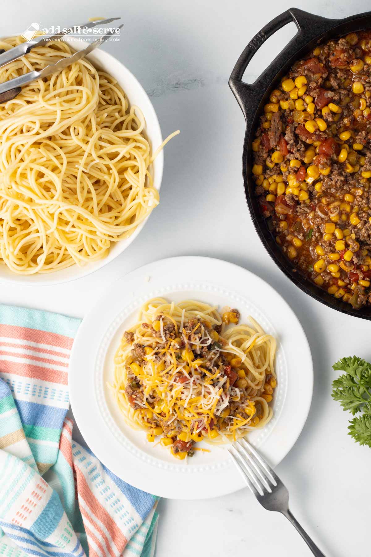 Plate of Mexican Spaghetti topped with shredded cheese on a counter beside a bowl of cooked spaghetti and a pot of Mexican Spaghetti sauce.