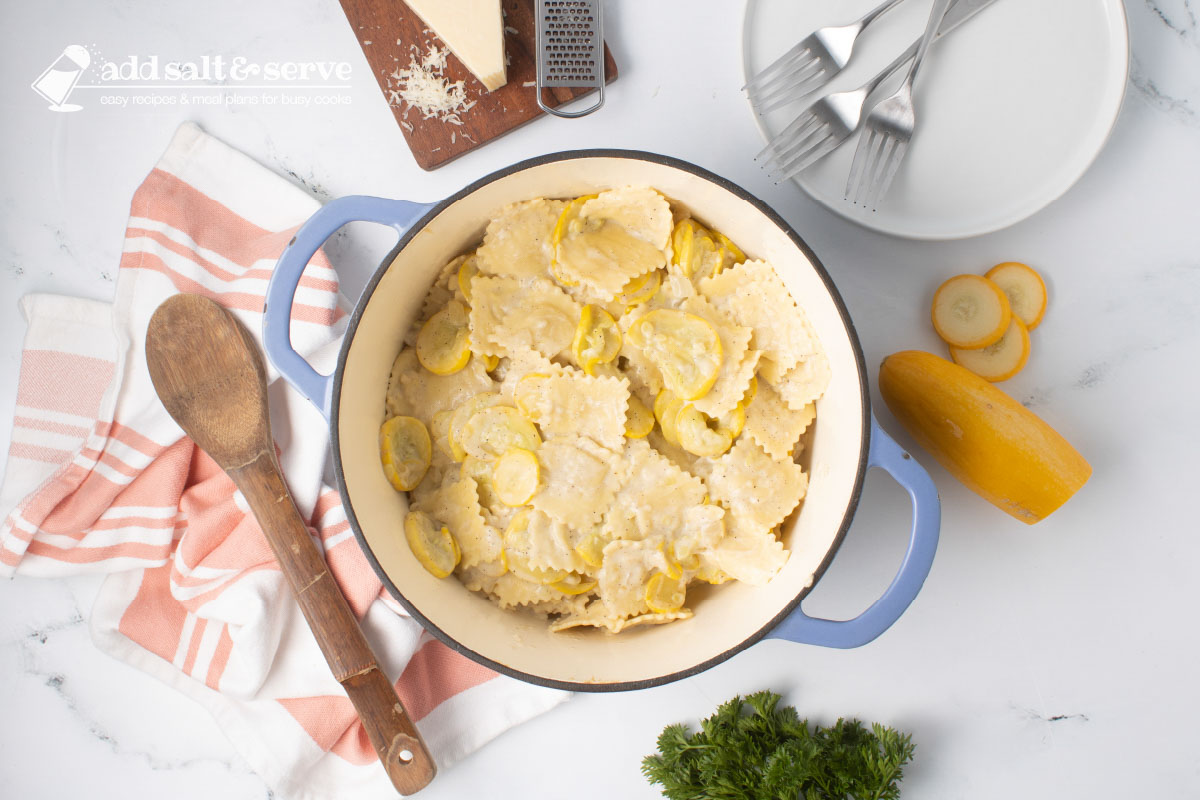 Dutch oven with cooked cheese ravioli mixed with Parmesan cheese and sautéed summer squash and onion beside a wooden spoon and surrounded by fresh squash, a bunch of parsley, and a block of fresh Parmesan cheese.