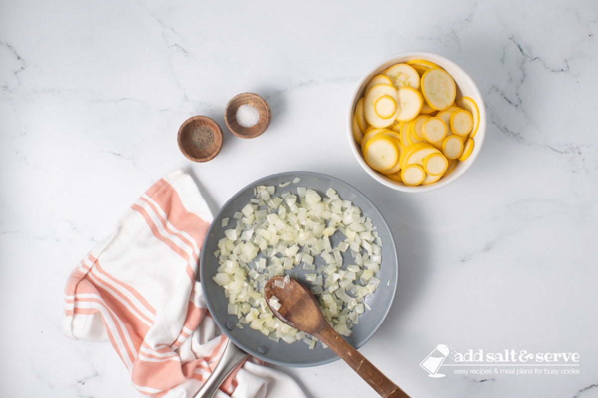Chopped onion being sautéed in a skillet with a wooden spoon beside a bowl of sliced yellow squash and small bowls of salt and pepper.