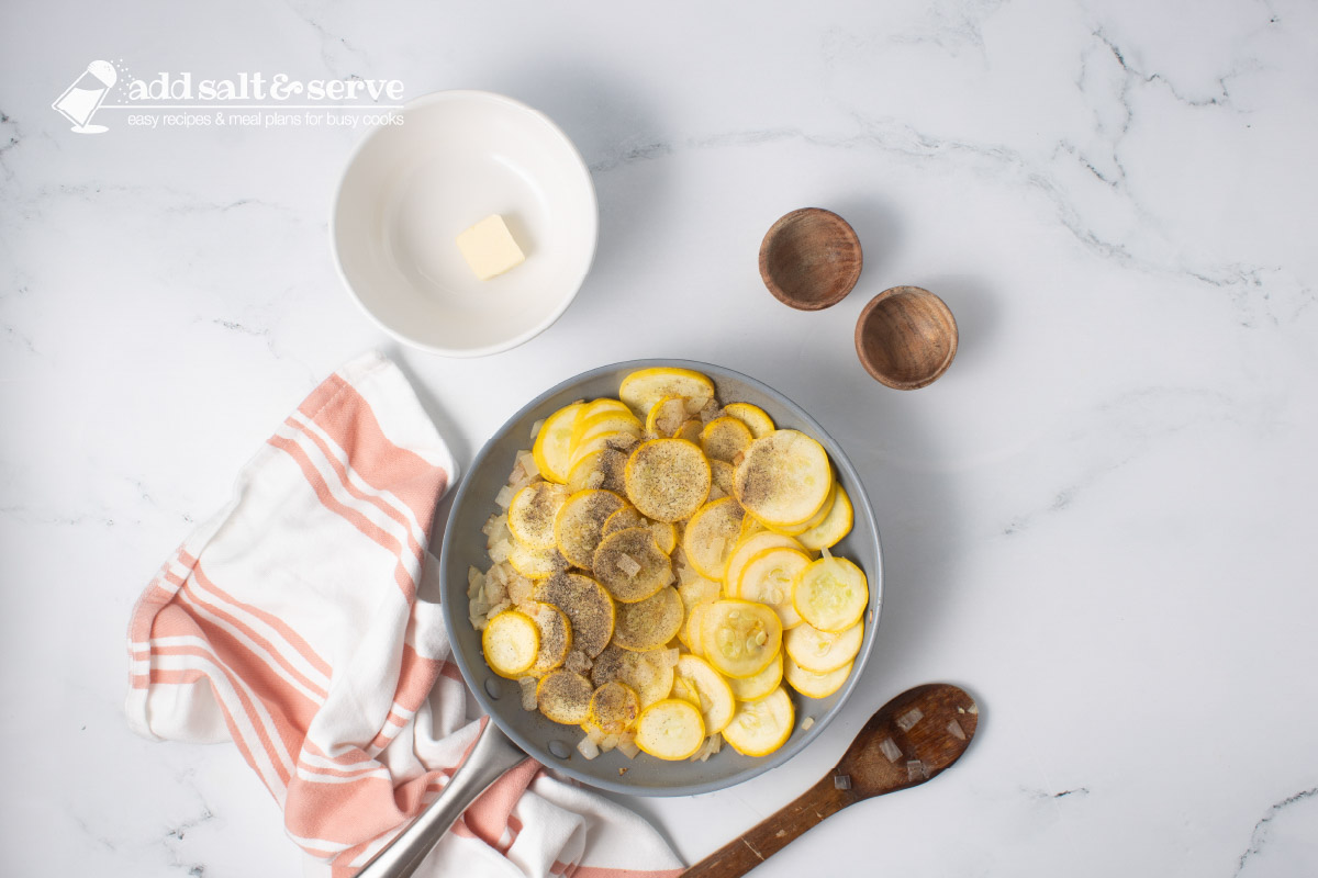 Yellow squash, chopped onion, salt, and pepper in a skillet with a wooden spoon and a bowl with a pat of butter.