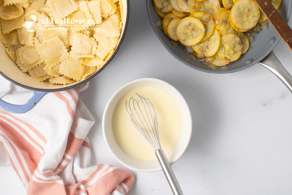 A bowl with melted butter, milk, and flour mixed with a whisk, a pot of cooked cheese ravioli, and a skillet of sautéed yellow squash and onion.