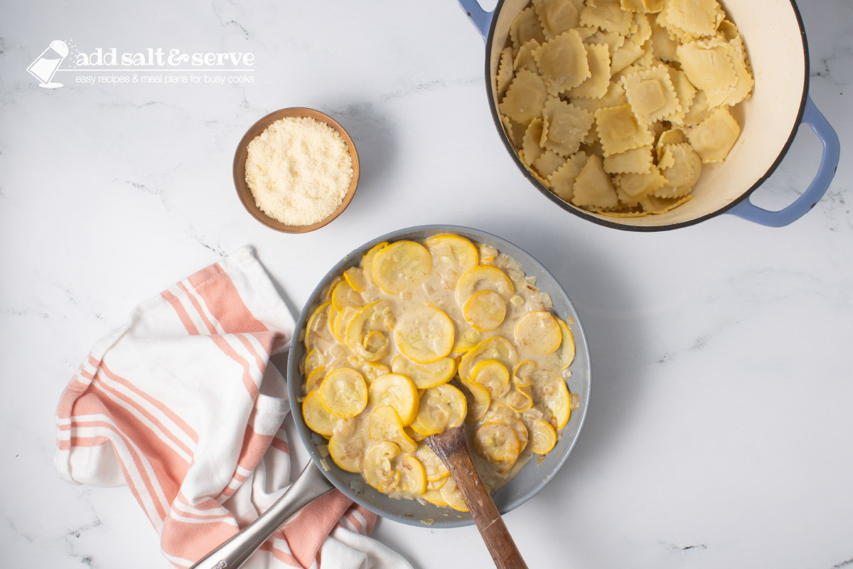 Skillet of sautéed yellow squash and onion after cooking with mixture of milk, flour, and butter beside a pot of cooked cheese ravioli and a small bowl of Parmesan cheese.