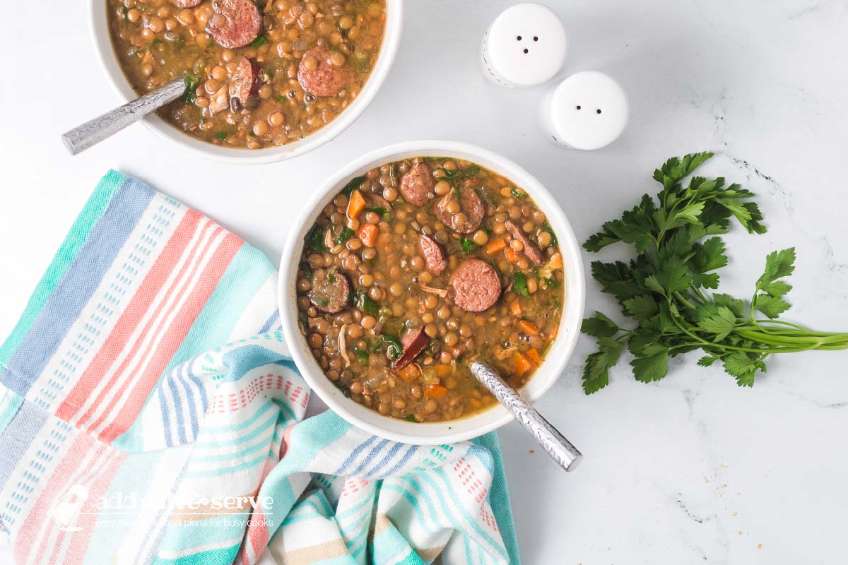 Bowl of lentil soup with kielbasa, spinach, carrots, onion, and green pepper beside a bunch of parsley and a tea towel.