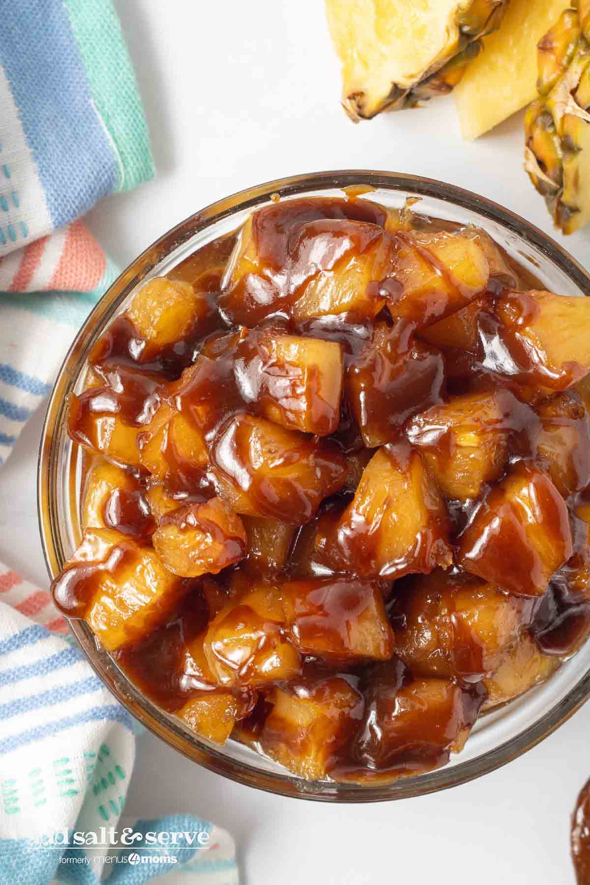 Bowl of caramelized pineapple chunks beside a tea towel and sliced pineapple on a counter.