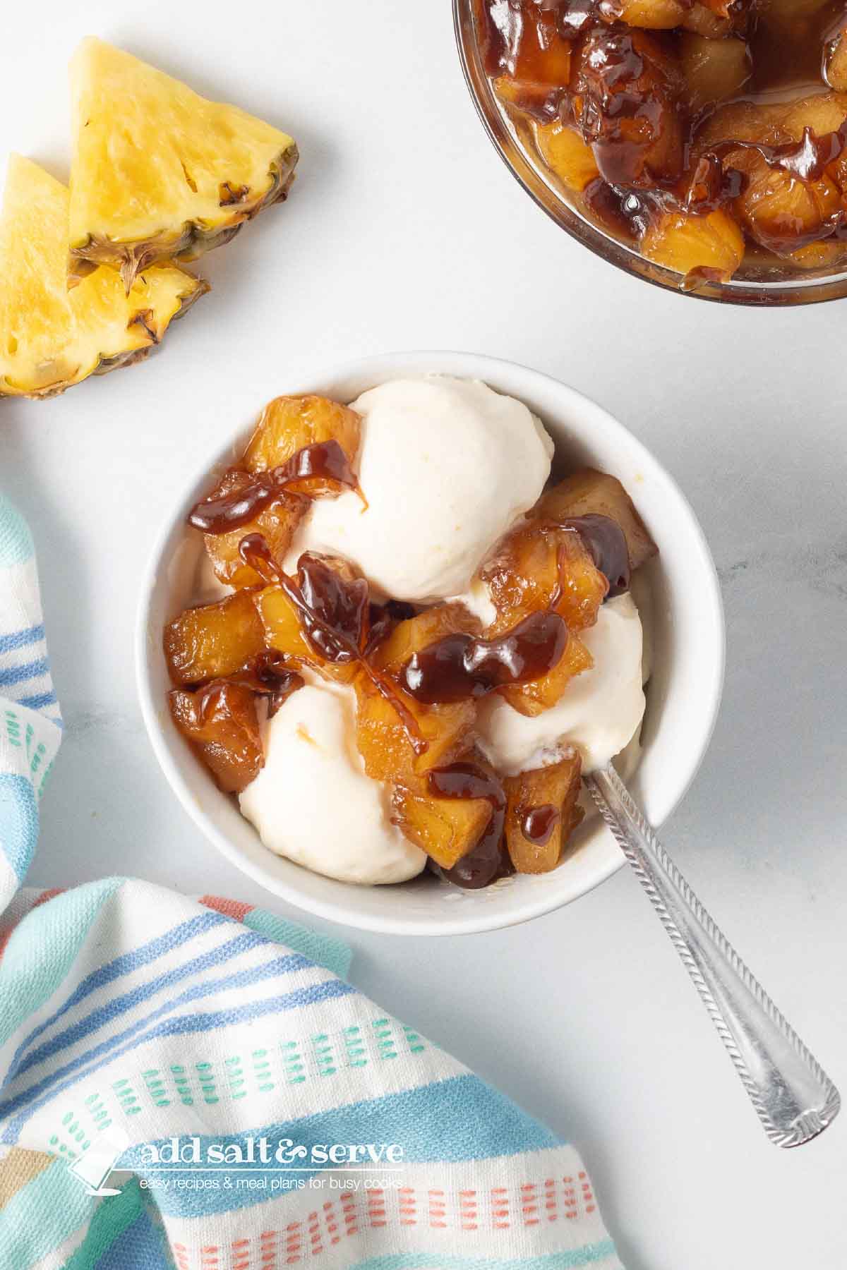 Bowl of caramelized pineapple chunks and vanilla ice cream drizzled with caramel sauce beside a tea towel and sliced pineapple on a counter.