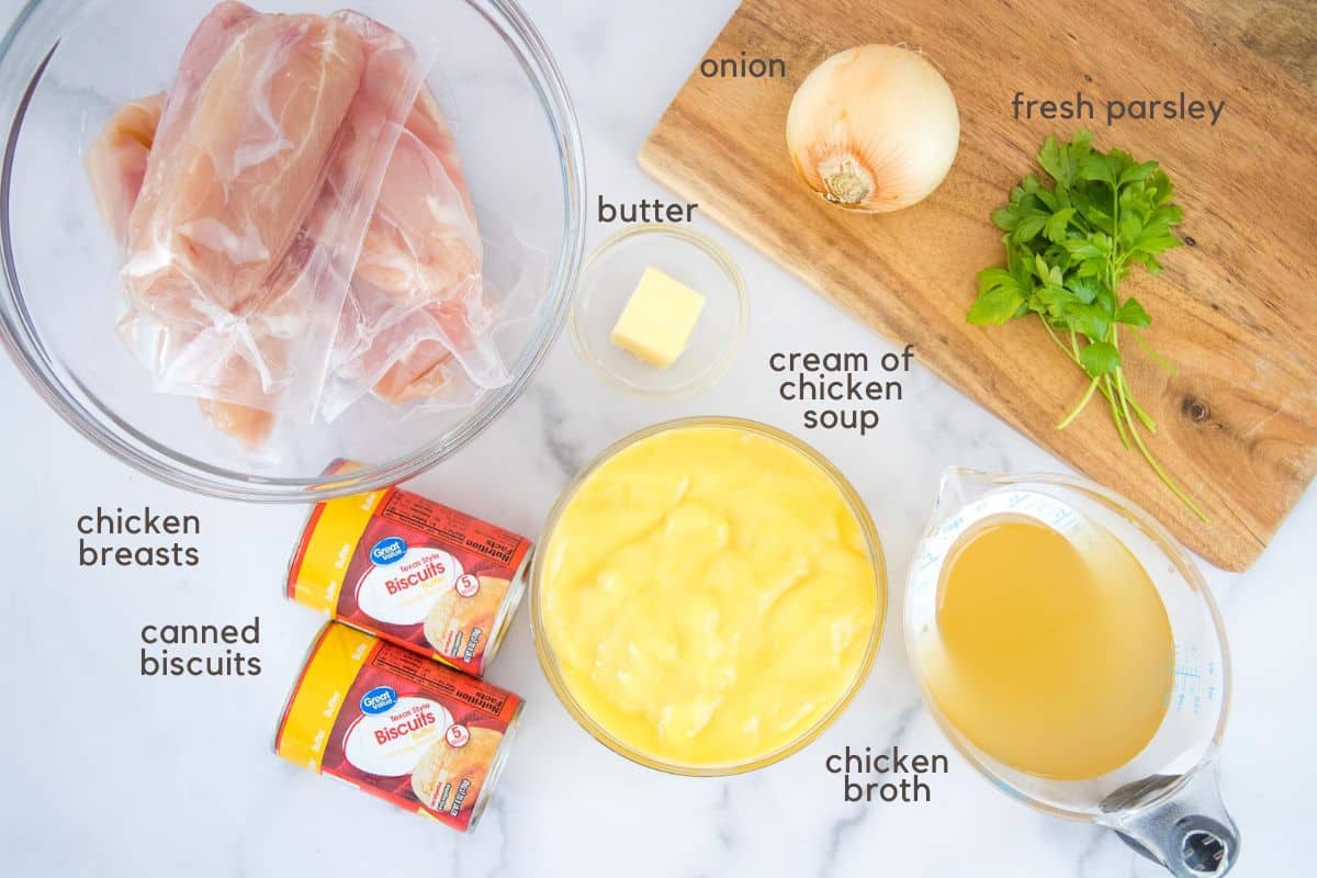 Individually wrapped chicken breasts in a clear glass bowl, two tablespoon butter in a small clear glass bowl, two cans of refrigerated biscuits, cream of chicken soup in a clear glass bowl, chicken broth in a clear measuring cup, and one onion and some sprigs of parsley on a cutting board.