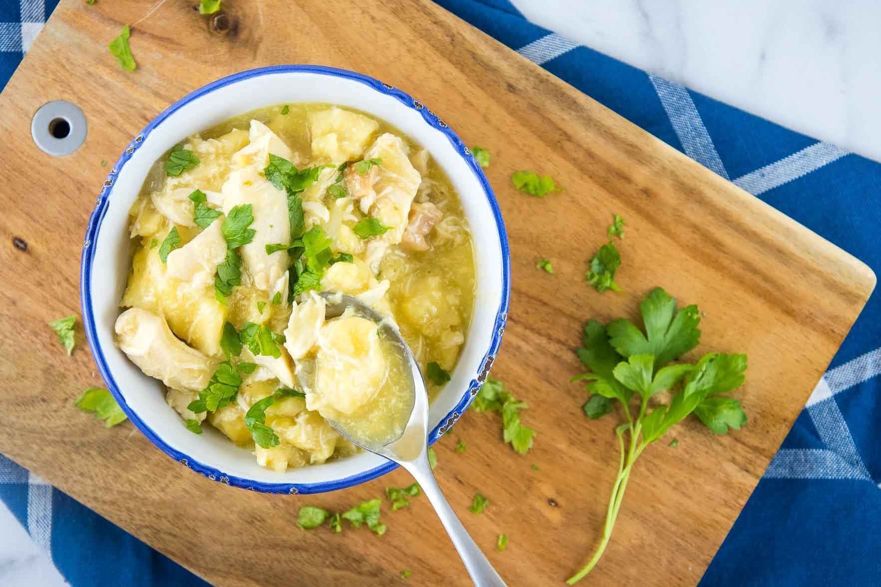 Chicken and dumplings garnished with parsley in a white bowl with a blue rim. There is a spoon with a dumpling and some broth. The bowl is on a wooden cutting board on top of a blue napkin with white stripes, all on a white marble counter.