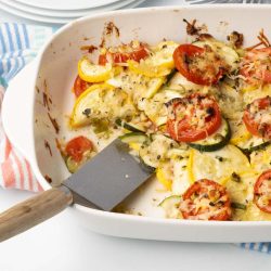 Baked layered casserole of yellow squash, zucchini, and tomatoes with herbs and cheese in a baking dish with a serving removed.
