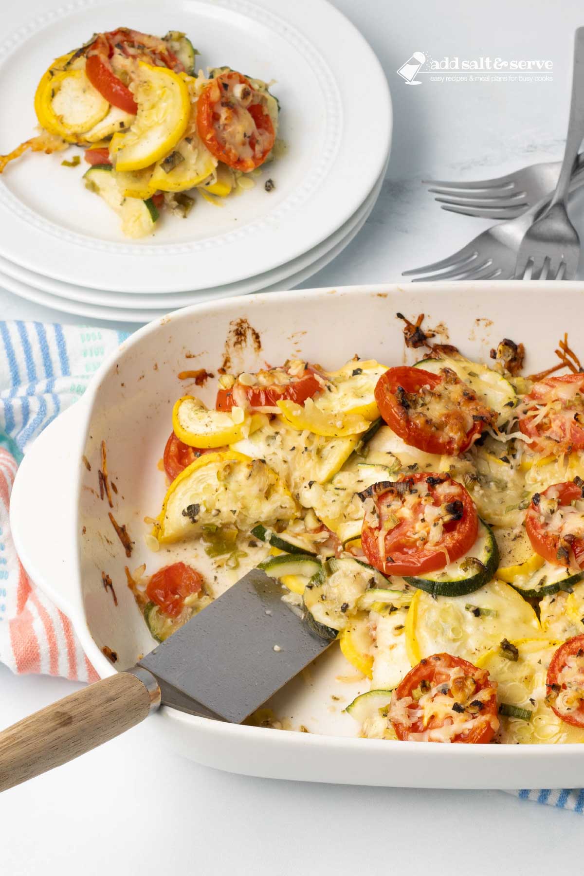 Baked layered casserole of yellow squash, zucchini, and tomatoes with herbs and cheese in a baking dish with a serving on a plate beside the dish.