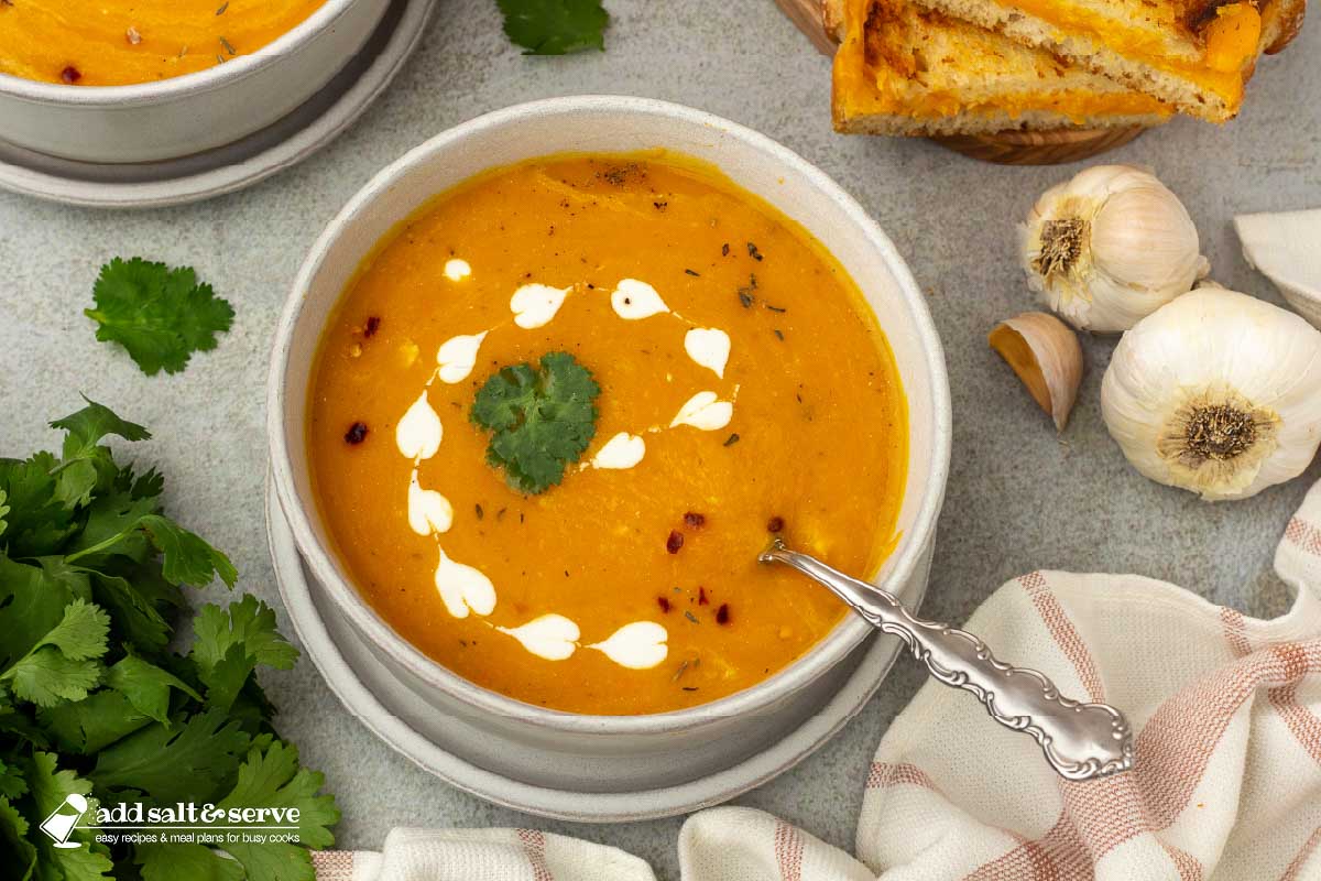 Bowls of carrot sweet potato soup beside a cutting board with grilled cheese sandwiches with garlic bulbs and parsley to the side.