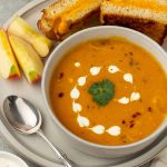 Bowl of carrot sweet potato soup on a plate with apple slices and a grilled cheese sandwich.