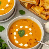 Bowls of carrot sweet potato soup beside a cutting board with grilled cheese sandwiches.