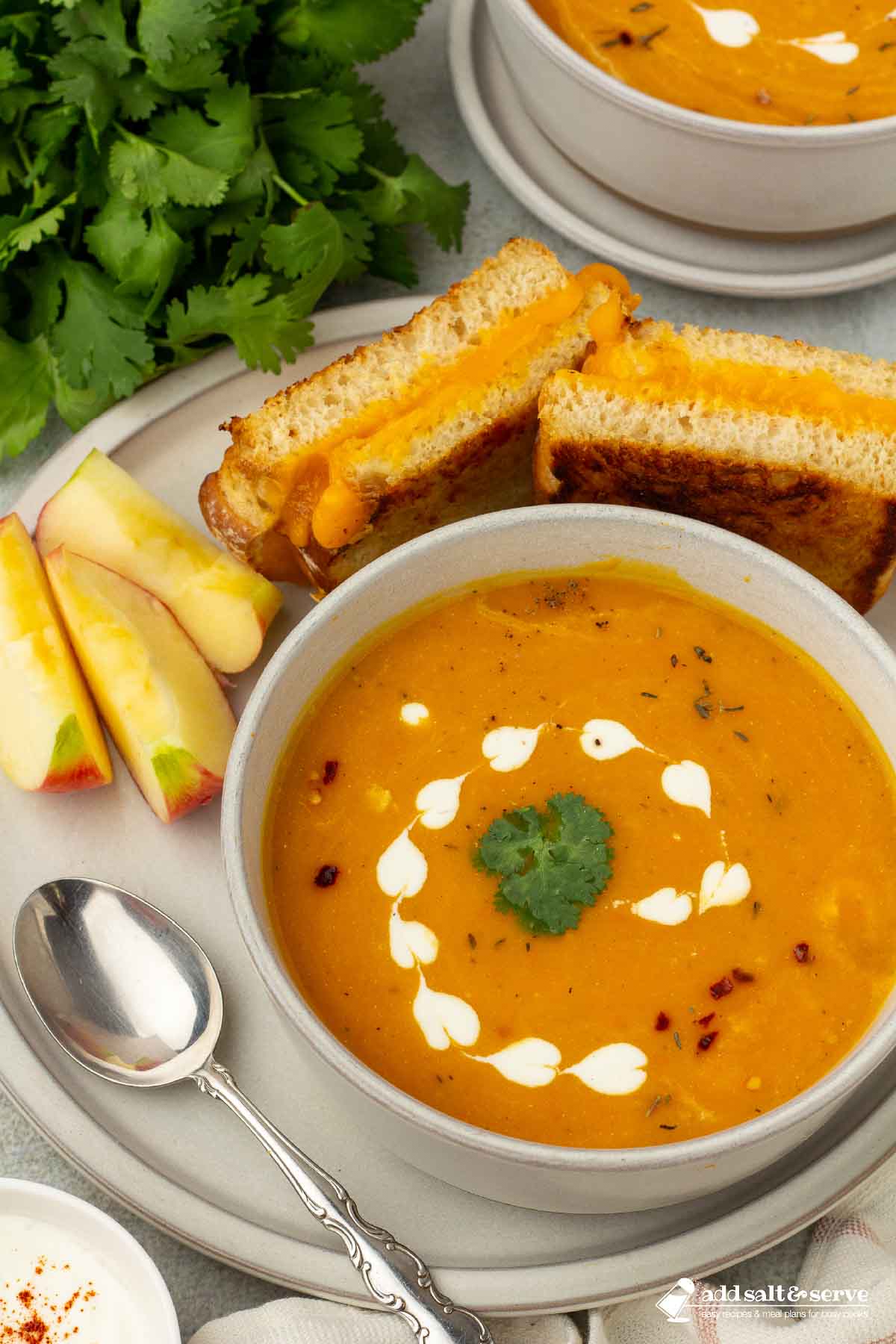 Bowl of carrot sweet potato soup on a plate with apple slices and a grilled cheese sandwich.