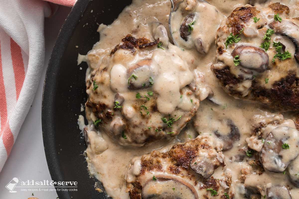 Several Salisbury Steak patties in mushroom cream sauce in a black pan.