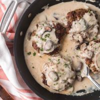 Several Salisbury Steak patties in mushroom cream sauce in a black pan with a serving spoon.
