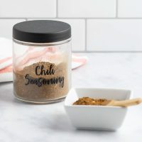 A jar of homemade chili seasoning beside a dish of the seasoning with a small wooden spoon.