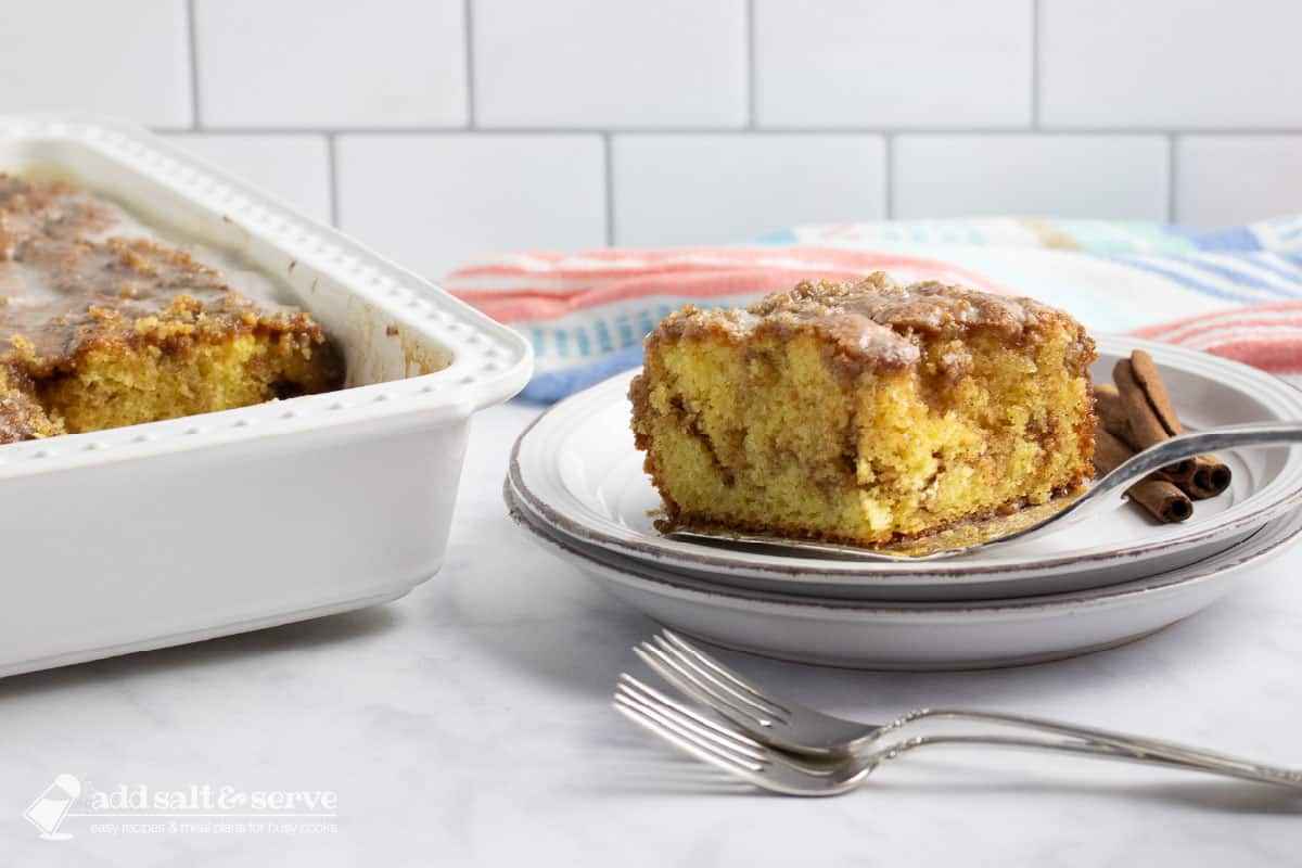 Slice of Honey Bun Cake being placed on a plate showing the cinnamon swirl in the middle and vanilla glaze on top on a counter beside the pan of cake and two forks.