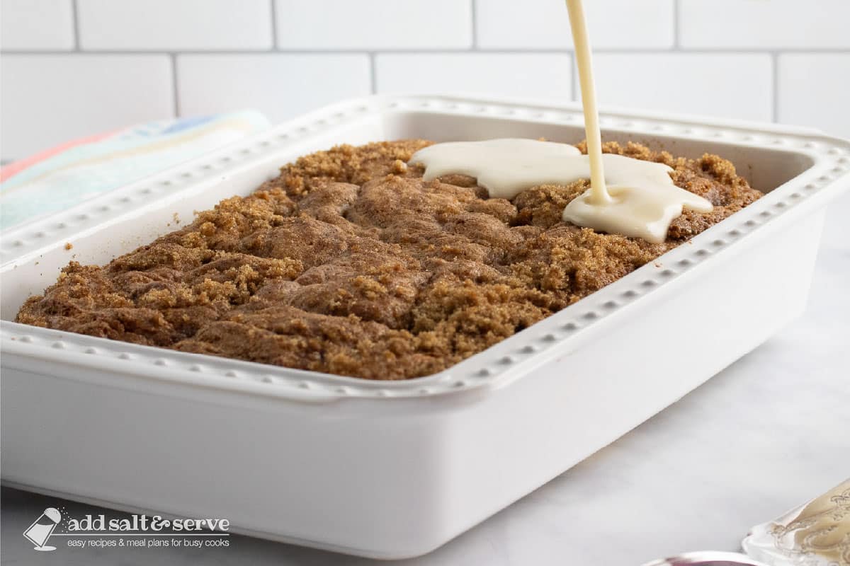 Baking pan with Honey Bun Cake with streusel topping with vanilla glaze being poured over the top.