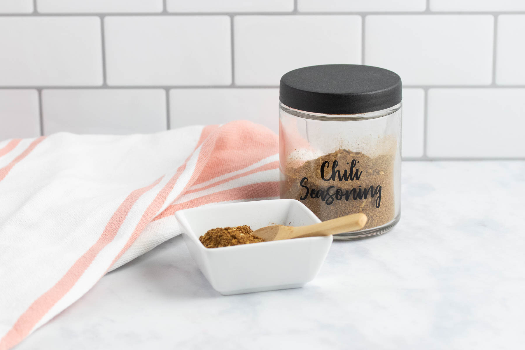 Jar of homemade chili seasoning beside a dish of the seasoning with a small wooden spoon.