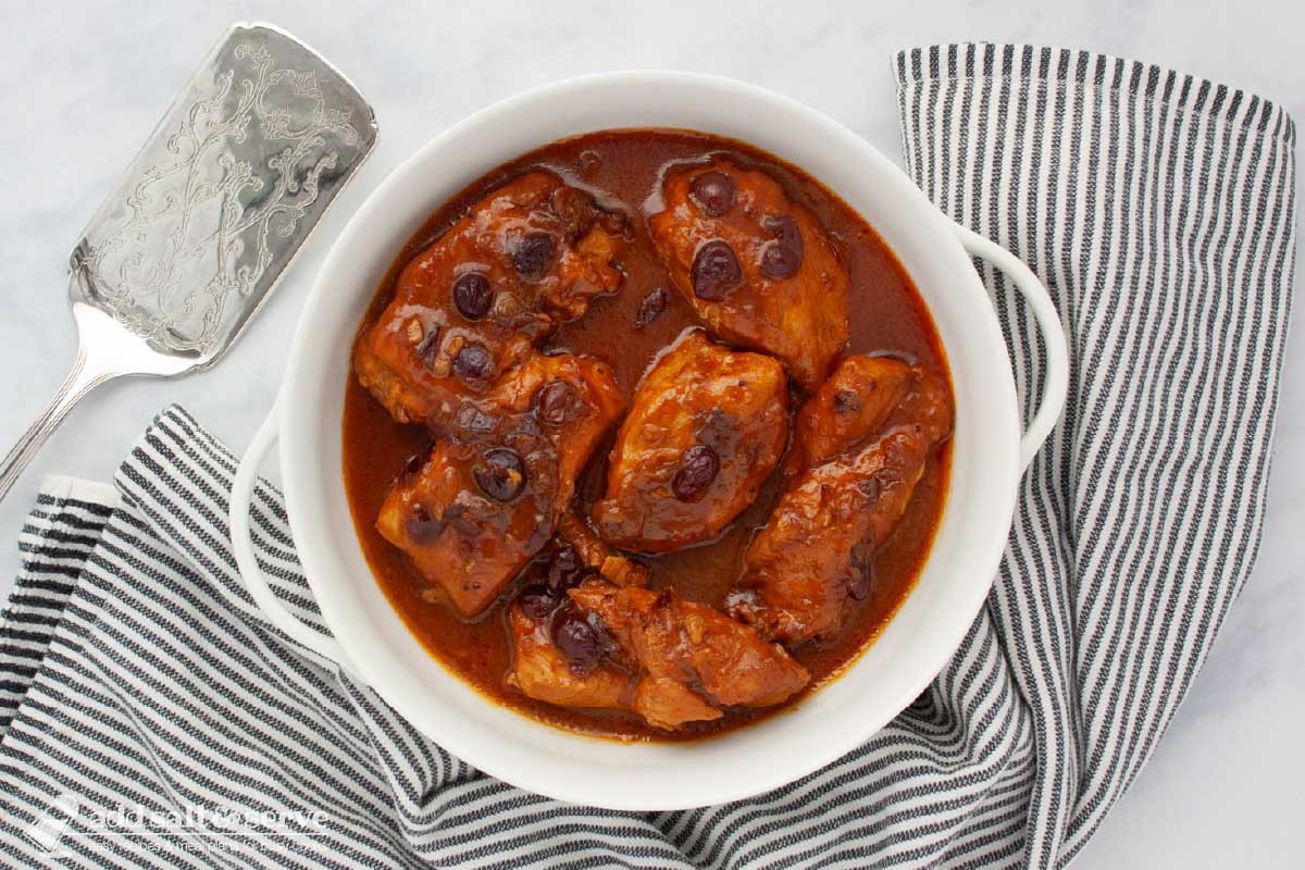 French Cranberry Chicken White serving dish with cooked chicken breasts in a sauce made from cranberry sauce with whole cranberries, French dressing, and onion soup mix beside a grey striped tea towel and a silver spatula.