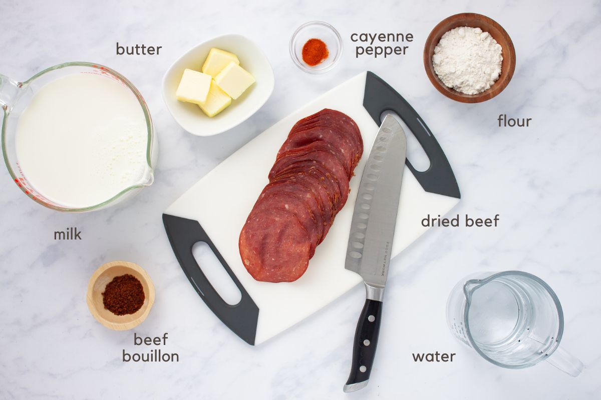 Cutting board with slices of dried beef and a knife, small bowls of flour, beef bouillon, cayenne pepper, and butter, and measuring cups with milk and water.