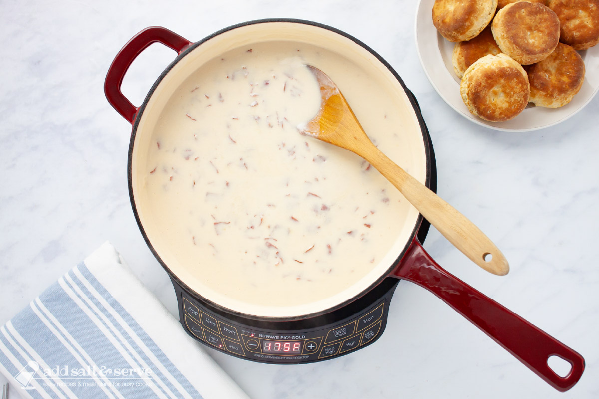Skillet of creamy chipped beef gravy with a wooden spoon beside a plate of biscuits.
