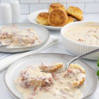Plate of creamy chipped beef gravy over two biscuit halves with a forkful held above the plate on a counter next to another plate of biscuits and gravy, a bowl of the gravy, and a plate of biscuits.