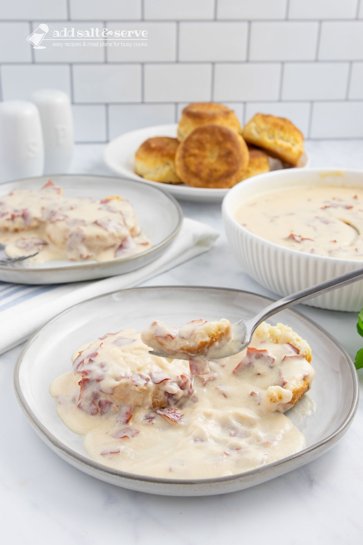 Plate of creamy chipped beef gravy over two biscuit halves with a forkful held above the plate on a counter next to another plate of biscuits and gravy, a bowl of the gravy, and a plate of biscuits.