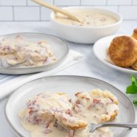 Plate of creamy chipped beef gravy over two biscuit halves with a fork on a counter next to another plate of biscuits and gravy, a bowl of the gravy, and a plate of biscuits.