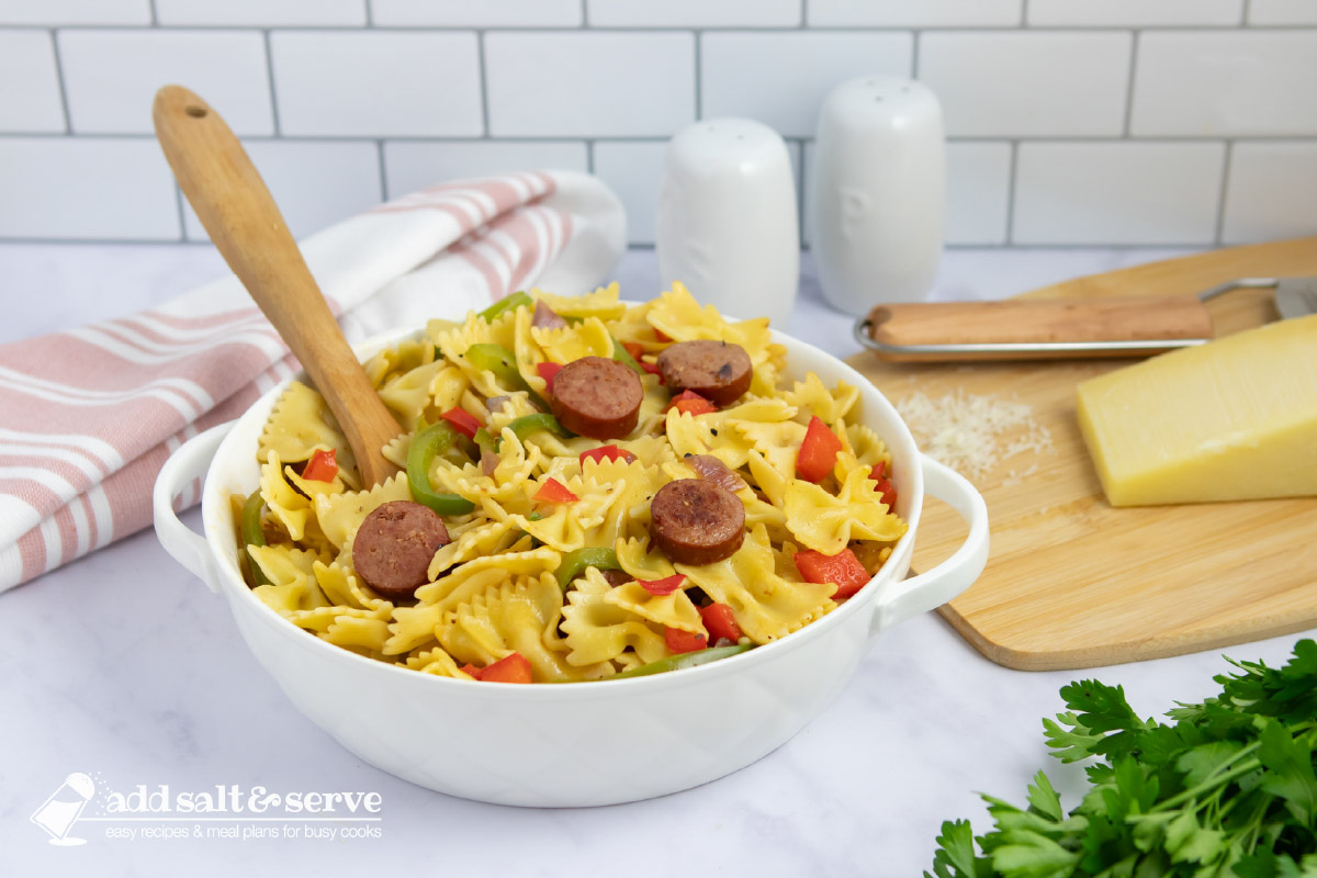 Bowtie pasta with red and green bell peppers, red onion, and sliced kielbasa with a spoon in a serving dish beside fresh parsley and Parmesan cheese.