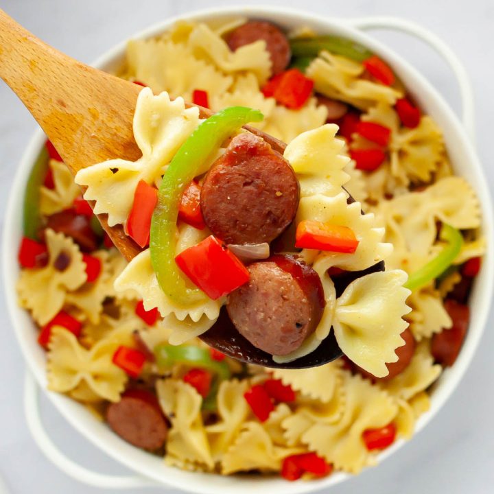 A wooden spoonful of bowtie pasta with red and green bell peppers, red onion, and sliced kielbasa held above a service dish of the same.