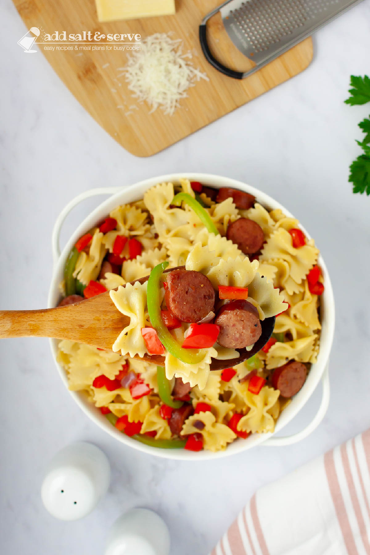 Bowtie pasta with red and green bell peppers, red onion, and sliced kielbasa with a spoon in a serving dish beside fresh parsley and Parmesan cheese.