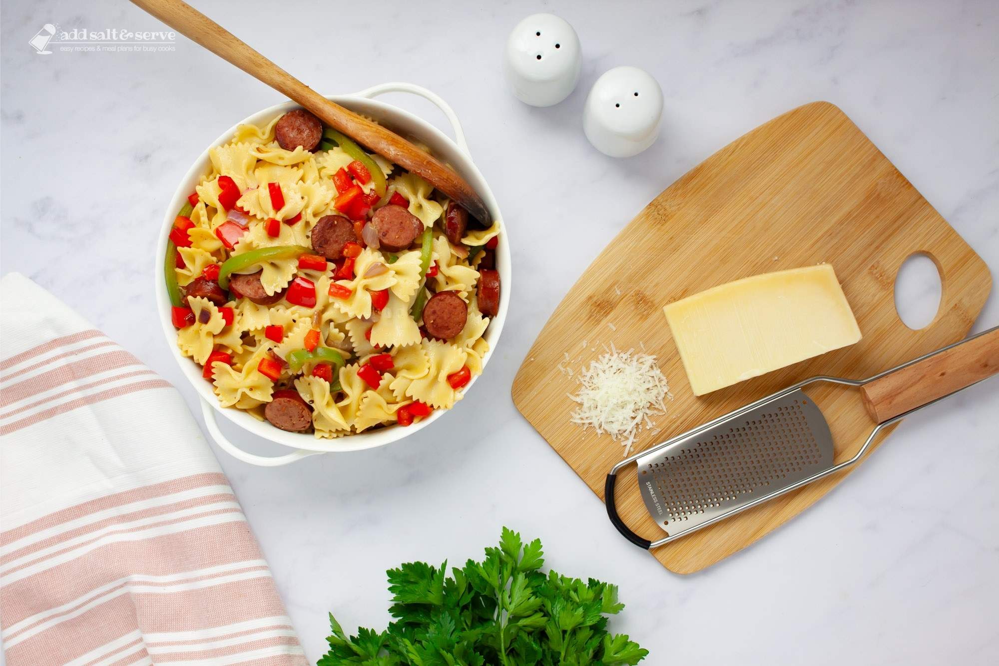 Bowtie pasta with red and green bell peppers, red onion, and sliced kielbasa with a spoon in a serving dish beside fresh parsley and Parmesan cheese.