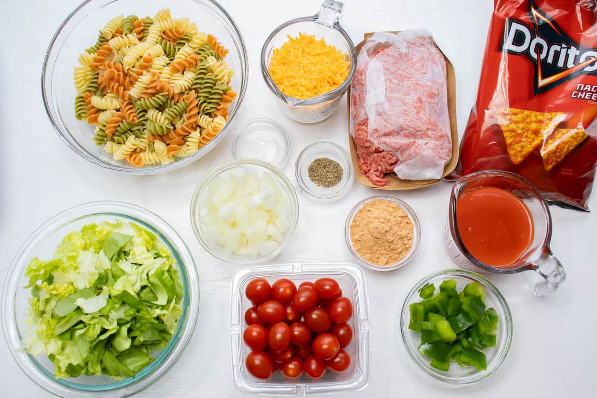 Overhead shot of ingredients cooked spiral pasta, lettuce, chopped onion, cherry tomatoes, chopped green bell pepper, taco seasoning mix, salt, pepper, shredded cheddar cheese, ground beef, French dressing, and nacho cheese Doritos.